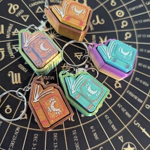 Puede incluir: Una colección de llaveros coloridos con forma de libros abiertos, cada uno con un diseño de media luna. Los llaveros son de varios tonos de naranja, verde, azul y morado, con llaveros plateados. El fondo es una superficie negra con símbolos astrológicos dorados.