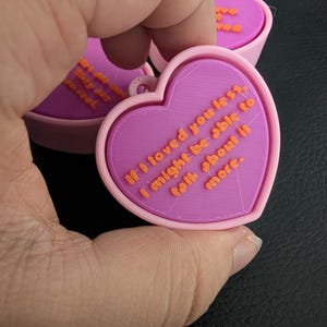 Könnte beinhalten: Ein herzförmiger Anhänger in Rosa und Lila mit dem Zitat "If I loved you less, I might be able to talk about it more" in orangefarbenen Buchstaben. Der Anhänger hat eine kleine Schlaufe zur Befestigung.