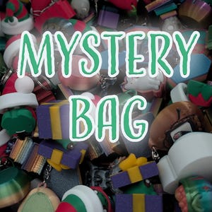 Könnte beinhalten: Eine Sammlung bunter Schlüsselanhänger und Schmuckstücke, darunter Weihnachtsmützen, Geschenkboxen und andere festliche Formen. Die Worte "MYSTERY BAG" sind grün mit weißer Umrandung dargestellt, was auf eine Überraschungsauswahl hindeutet.