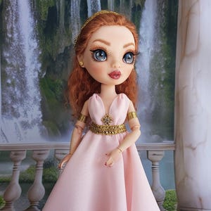 May include: A doll with long wavy auburn hair and blue eyes wearing a floor-length pale pink sleeveless gown. The dress features a gold metallic waistband with a central crystal embellishment, matching gold arm cuffs, and a gold headpiece.