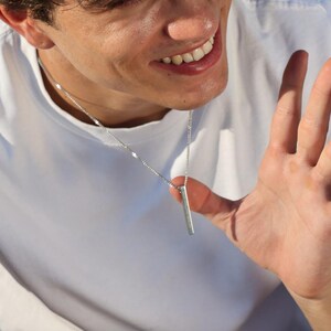 May include: A young person with curly brown hair is sitting on a blue and white surface. They are wearing a white t-shirt with a silver necklace and a black pair of shorts. They are smiling and waving with their right hand.