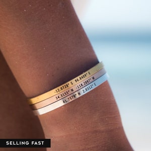 May include: Three gold, silver, and rose gold bracelets with coordinates engraved on them. The bracelets are stacked on a person's wrist. The coordinates are: -12.0728 S 96.8409 E, 34.3155 N-118.2097" W, 41.7579 N 2.0312" E.