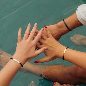 May include: Three people with their hands clasped together in a pool. They are wearing black and gold bracelets.