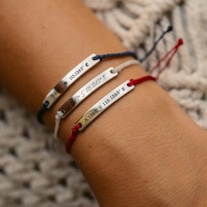 Può includere: Tre braccialetti con ciondoli argentati al polso di una persona. I ciondoli sono incisi con coordinate. I braccialetti sono rosso, bianco e blu.