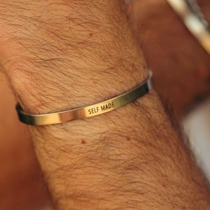 May include: A gold metal bracelet with the words "SELF MADE" engraved on it.