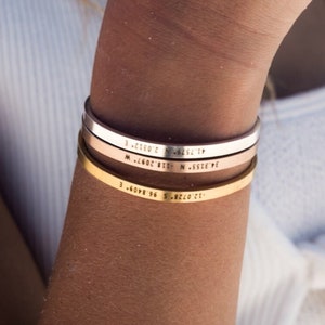 May include: Three gold, silver, and rose gold bracelets stacked on a person's wrist. Each bracelet has a different inscription.