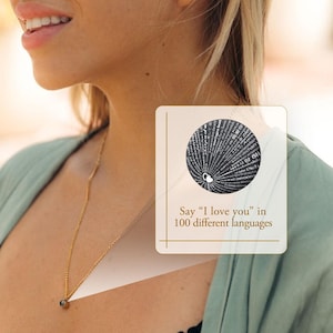 May include: A gold chain necklace with a small, round, dark gray pendant. The pendant has the words "I love you" written in 100 different languages.