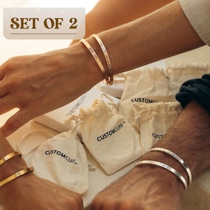 May include: Set of two gold and silver engraved bracelets with the text "CUSTOMCUFF" on the pouches they are in. The bracelets are on the wrists of two people.