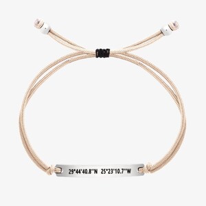 Puede incluir: Un brazalete de cordón beige con una etiqueta plateada que dice "29°44'40.8"N 25°23'10.7"W".
