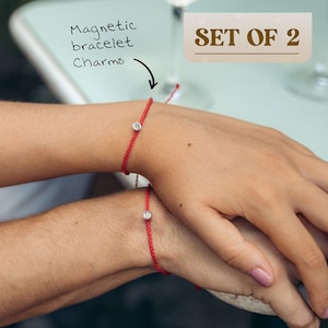 May include: Two red string bracelets with small silver magnetic charms. The bracelets are worn on the wrists of two people. The text "SET OF 2" is visible in the image.