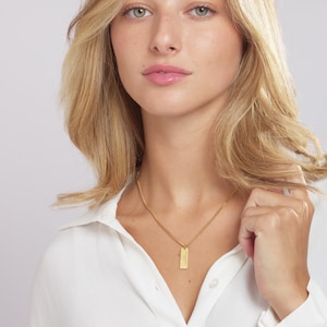 May include: A woman wearing a white shirt with a gold chain necklace and a small gold bar pendant.