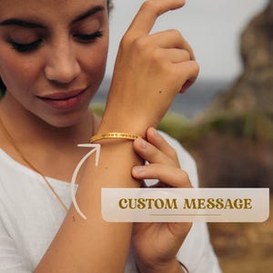 May include: A gold-toned bangle bracelet with a custom message engraved on it. The bracelet is worn on a person's wrist.