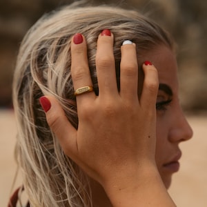 May include: A gold ring with the inscription "remarkable" on a woman's finger. The ring is on the middle finger of her right hand. Her nails are painted red.