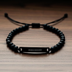 May include: Black beaded bracelet with a rectangular plate engraved with the Roman numerals XI·XIX·MMXXIV. The bracelet is made of small, round, dark beads and adjustable black cords. The bracelet is displayed on a wooden surface.