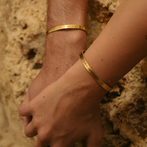 May include: Two gold bracelets on wrists. One bracelet has the coordinates "47.7298°N - 122.0715°W" engraved on it.