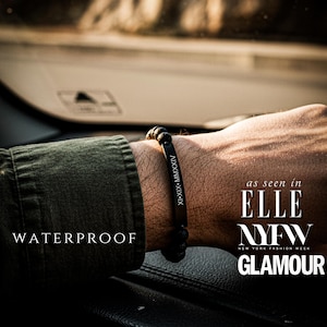 Puede incluir: Una pulsera de cuentas negras con los números romanos XI-XIX-MMXXIV grabados en una placa de metal negro. La palabra "WATERPROOF" se muestra en texto blanco. La pulsera aparece en ELLE NYFW GLAMOUR.