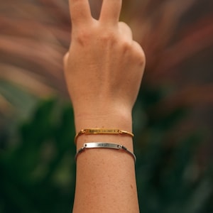 May include: Two gold and silver bracelets on a wrist. The gold bracelet has the coordinates "6.1885° S 116.1583° E" engraved on it. The silver bracelet has "JN X 1.18" engraved on it.