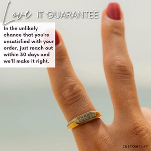 Puede incluir: Un anillo de oro con una inscripci&oacute;n personalizada que dice "52 36 46.08 N 4.91 13.91 E". El anillo est&aacute; en un dedo, y el fondo es una imagen borrosa de una playa.