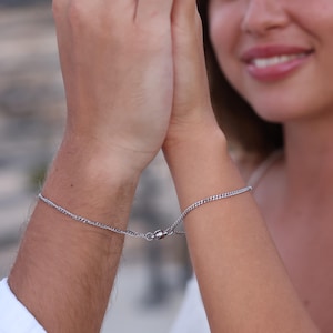 May include: Two people wearing matching silver chain bracelets connected by a magnetic clasp. The bracelets are on their wrists, and their hands are touching.