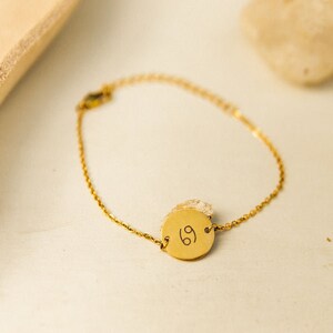 May include: A gold chain bracelet with a round charm featuring the Cancer zodiac symbol.