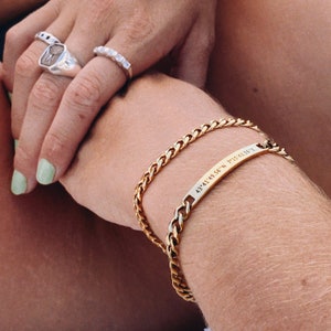 Puede incluir: Un brazalete de cadena dorada con un dije de barra dorada grabado con las coordenadas "43.4149.56'N 71.543.55'E".