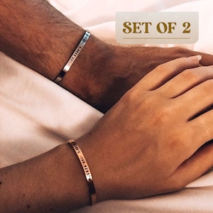 May include: Set of two gold-toned bracelets with engraved text. The bracelets are on the wrists of two people holding hands.