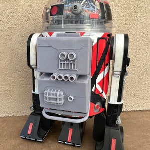 May include: A red, white, and black robot toy with a clear plastic dome head. The robot has a gray panel with a series of holes and a grill on the front.
