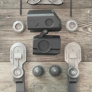 Puede incluir: Un juguete robot desmontado con piezas de pl&aacute;stico gris y negro. El robot tiene dos piernas, un cuerpo, una cabeza y dos antenas. Las piezas est&aacute;n dispuestas sobre una superficie de madera.