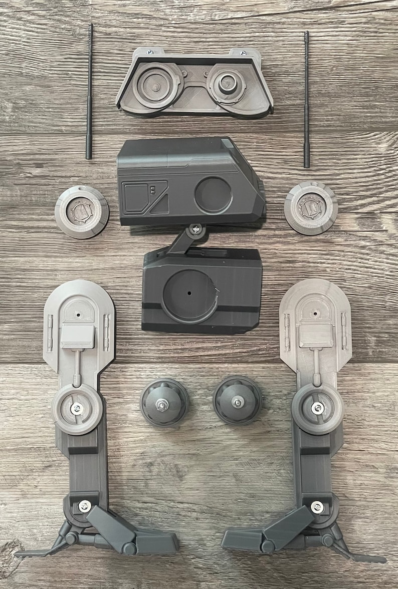 Puede incluir: Un juguete robot desmontado con piezas de pl&aacute;stico gris y negro. El robot tiene dos piernas, un cuerpo, una cabeza y dos antenas. Las piezas est&aacute;n dispuestas sobre una superficie de madera.