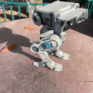 Puede incluir: Un juguete robot blanco y gris con detalles azules. El robot tiene dos antenas y est&aacute; de pie sobre una superficie gris.