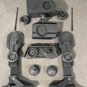 Puede incluir: Un modelo de robot impreso en 3D negro desmontado con m&uacute;ltiples partes, incluyendo una cabeza, piernas y ruedas.