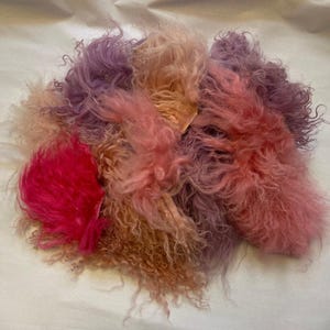 Puede incluir: Un montón de fibras coloridas y esponjosas en tonos rosa, morado y melocotón. La textura parece suave y rizada, con una sección fucsia vibrante a la izquierda. Las fibras están dispuestas sobre una superficie blanca.