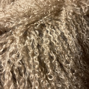 May include: Close-up of a beige, curly textured fabric. The fabric has a dense, looped pile, creating a soft, wavy appearance. The colour is a warm, neutral tone, with variations in light and shadow across the surface.