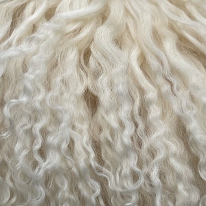 Puede incluir: Primer plano de una tela texturizada de color crema con fibras largas y onduladas. Las fibras tienen un aspecto suave y rizado, creando una textura afelpada y acogedora. El fondo es de color beige claro, realzando el tono cremoso de la tela. El efecto general es de calidez y confort.