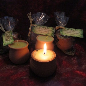 Puede incluir: Varios recipientes de terracota con velas encendidas y envueltos en celofán. Las velas son amarillas y los recipientes son pequeños y redondos. Cada recipiente tiene una etiqueta que dice "Yule Pot". El fondo es rojo oscuro.