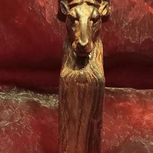 Op de afbeelding: Houten sculptuur van een ram met gedetailleerde hoorns en een getextureerd, verweerd uiterlijk. De ram is bruin met donkere accenten, tegen een rode achtergrond. De sculptuur heeft een rechthoekige basis.