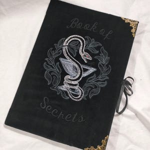 May include: Black book with the words "Book of Secrets" embroidered in script. The cover features an embroidered design of a snake coiled around a chalice within a wreath. Gold-coloured corner protectors and a black ribbon closure complete the design.