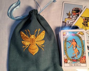 May include: A teal drawstring pouch featuring a gold embroidered bee on the front and light blue ribbon ties, positioned next to a deck of tarot cards with the card labeled THE WORLD visible on top, showing a figure inside a laurel wreath.