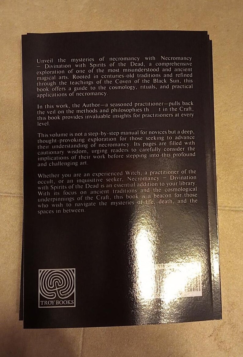 May include: A black book titled "Necromancy Divination with Spirits of the Dead" by Troy Books. The back cover has text about necromancy, rituals, and ancient traditions. The book's logo is a spiral design.