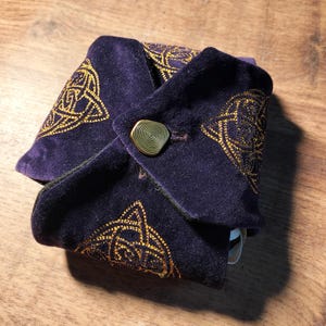 以下が含まれることがあります： 金色のケルトノット刺繍が施された濃い紫色のベルベットポーチ。ポーチは金のボタンで留められ、ユニークな折り畳みデザインが特徴です。ポーチは、小さなアイテムを保管するために使用される可能性があります。