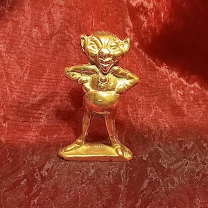 May include: A small, golden figurine of a goblin-like creature with arms akimbo. The figure has a wide grin, pointed ears, and a bulbous belly. It stands on a rectangular base, and the overall design is detailed and whimsical.