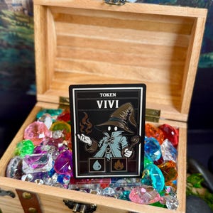Puede incluir: Un cofre del tesoro de madera lleno de gemas de colores. Dentro del cofre hay una ficha en blanco y negro con el nombre "VIVI" y una ilustración de un mago. La ficha tiene un sombrero de mago y un bastón.