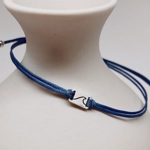 Puede incluir: Una pulsera de cordón azul con un pequeño dije plateado con un diseño de ola. La pulsera se exhibe sobre un jarrón de cerámica blanco, mostrando su diseño minimalista y su tema inspirado en el océano.