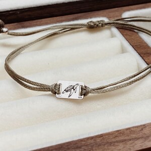 Könnte beinhalten: Olivgrünes Armband mit einem kleinen rechteckigen Anhänger mit einem Schildkröten-Design. Das Armband besteht aus einem dünnen, gewebten Kordel und hat verstellbare Gleitknoten. Der Anhänger ist silberfarben und das Armband wird in einer Holzschmuckschatulle präsentiert.
