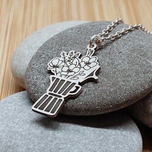 Puede incluir: Un collar de plata con un colgante que representa un ramo de flores en un jarrón. El colgante está grabado con un diseño floral detallado.