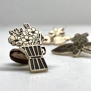 Può includere: Uno spillo di legno con un design in bianco e nero di una caffettiera con fiori. Lo spillo è su un chicco di caffè marrone.