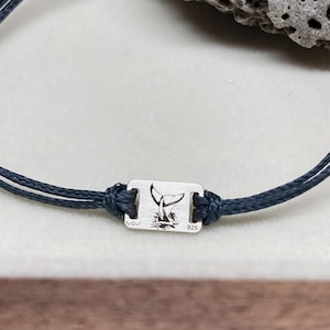 Può includere: Primo piano di un braccialetto blu navy con un ciondolo rettangolare in argento. Il ciondolo presenta un design a coda di balena e il numero "925". Il braccialetto è realizzato con un cordoncino intrecciato.