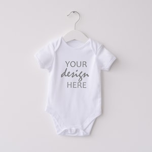 May include: White baby onesie with short sleeves, hanging on a white hanger. The text "YOUR design HERE" is printed in gray on the front of the onesie. The onesie is made of a soft material and is designed for infants.