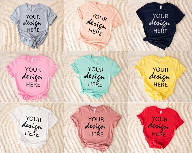 May include: Nine different colored t-shirts with the text "YOUR design HERE" printed on the front. The t-shirts are laid out on a white fluffy surface.