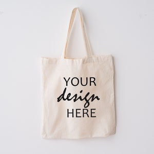 Könnte beinhalten: Eine schlichte weiße Canvas-Tragetasche mit schwarzem Text, der "YOUR design HERE" lautet.